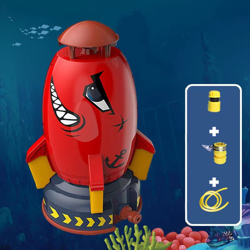 Red rocket water sprinkler toy with shark design, yellow fins, and hose connectors on a blue underwater background.