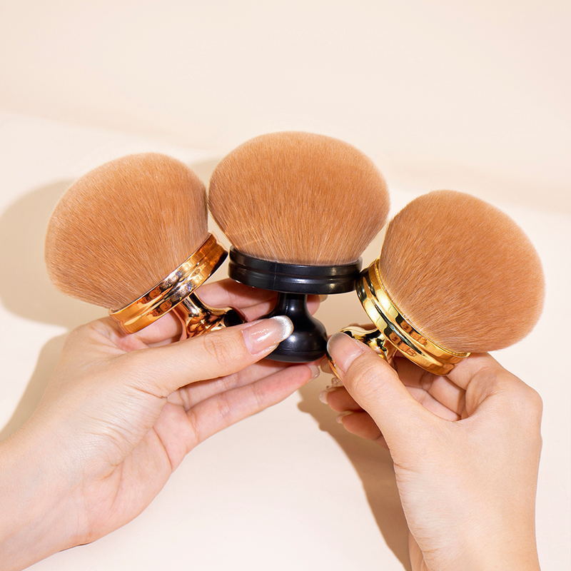 Hands hold three oversized, soft, rounded body powder brushes with metallic rose gold, black, and gold bases.