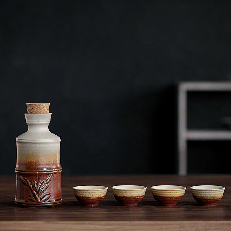 Ceramic sake set with bamboo-style carafe, cork stopper, and four matching gradient-brown cups on a dark wood table.