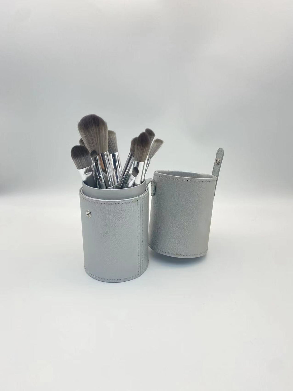 A set of soft-bristled makeup brushes with silver ferrules inside a matching grey cylindrical leather case.