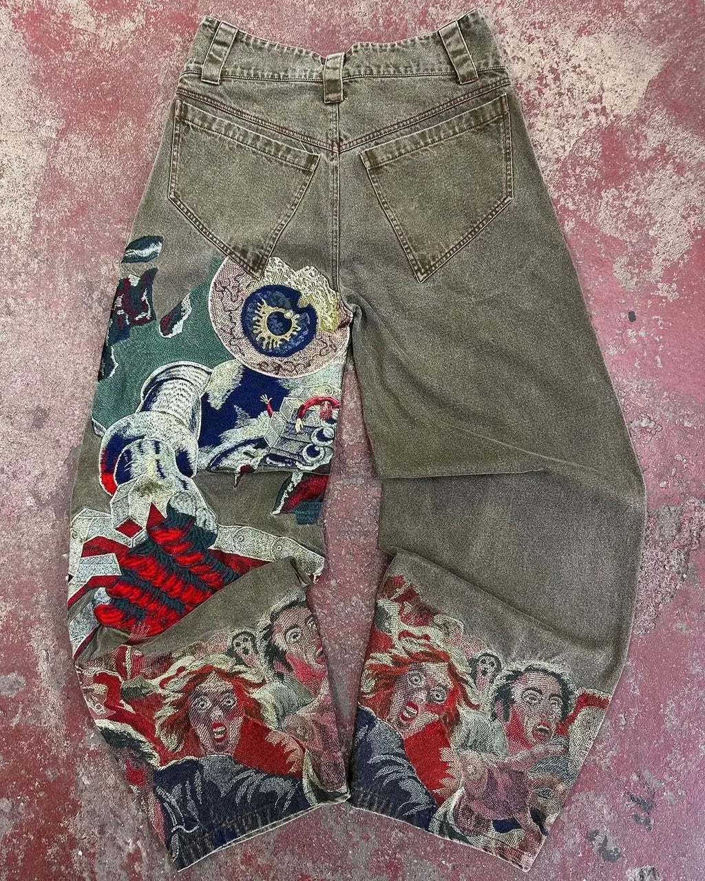 Washed grey baggy jeans featuring colorful artistic patchwork embroidery with surreal figures and vibrant designs.