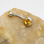 Curved titanium steel navel barbell with two sparkling yellow crystal gems resting on a beige stone.