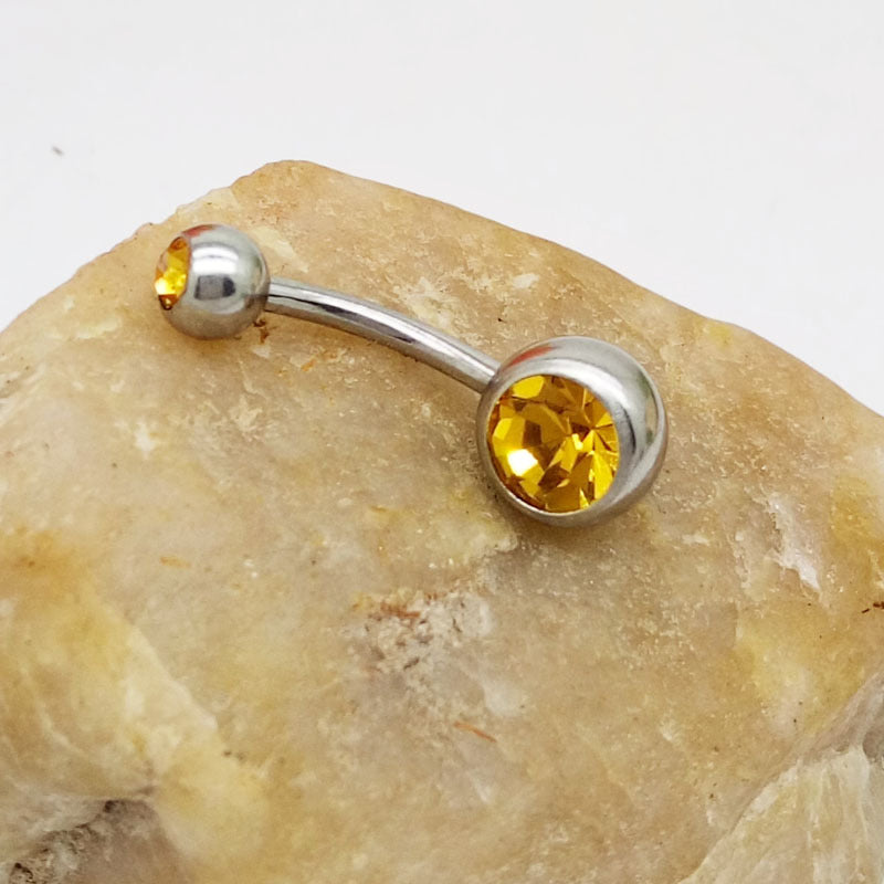Curved titanium steel navel barbell with two sparkling yellow crystal gems resting on a beige stone.