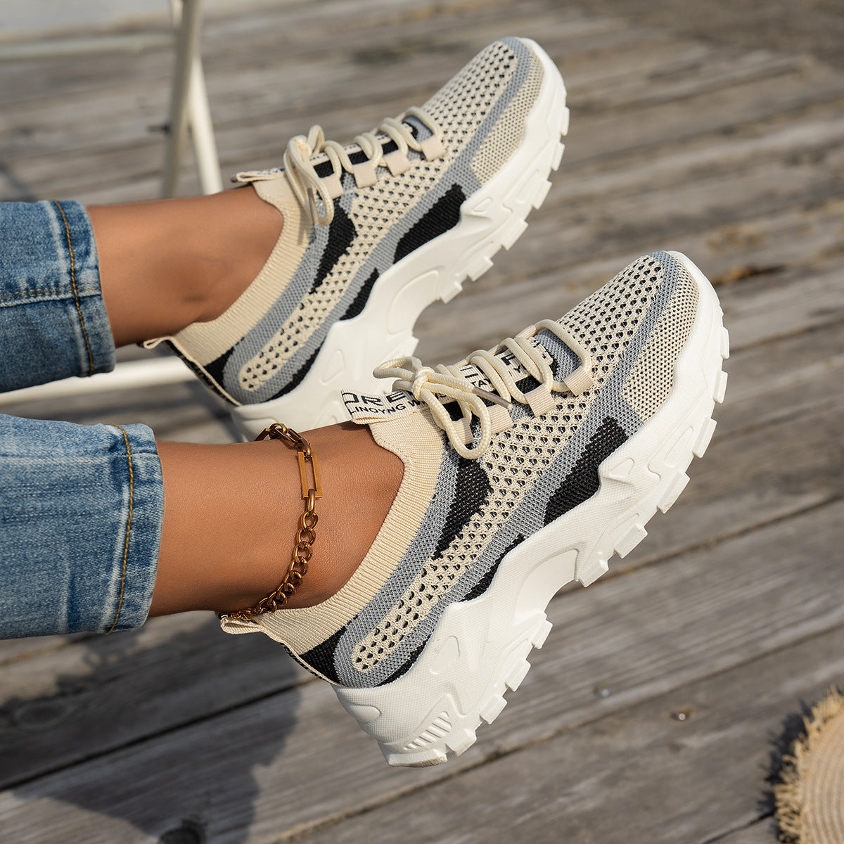 Beige and black mesh lace-up sneakers with thick white rubber soles, worn with blue jeans on a wooden surface.