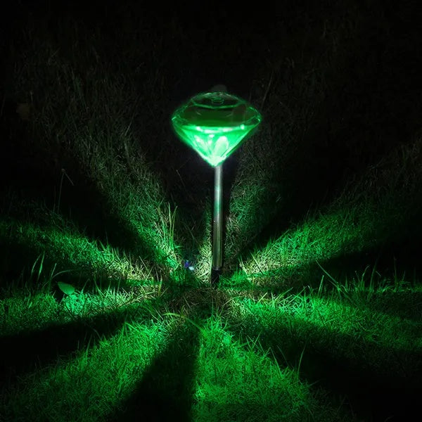 A diamond-shaped solar garden light with a silver stake, glowing bright green on dark grass.