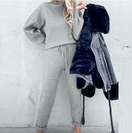 A person wears a grey long-sleeve fleece sweater suit with drawstring pants, holding a black faux-fur hooded jacket.