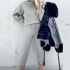A person wears a grey long-sleeve fleece sweater suit with drawstring pants, holding a black faux-fur hooded jacket.