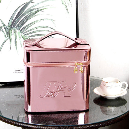Metallic rose gold cube-shaped makeup bag with handle, zipper closure, and embossed logo on a glass table.
