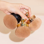 Hand holding three oversized, soft, tan-bristled makeup brushes with gold and black metallic handles.