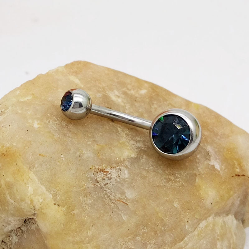 Silver titanium belly button ring with two dark blue faceted crystals resting on a piece of rough beige stone.