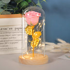 A glittery pink rose with gold leaves, illuminated by LED string lights inside a glass dome on a wooden base.