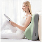 A woman reads while leaning against a contoured, two-tone sage green and grey fabric ergonomic wedge pillow on a bed.