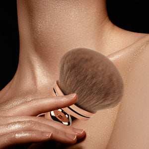 Oversized, soft brown powder brush with a rose gold handle, being used on glowing skin for neck and body application.