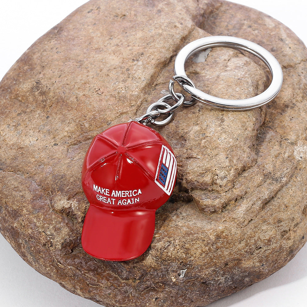 Red metal cap keychain with "MAKE AMERICA GREAT AGAIN" text and flag detail, on a silver ring atop a brown stone.