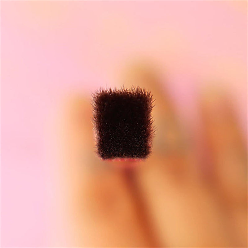 A close-up of a multi-purpose makeup brush with a dense, flat-top, squared-off dark brown bristle head on a pink background.