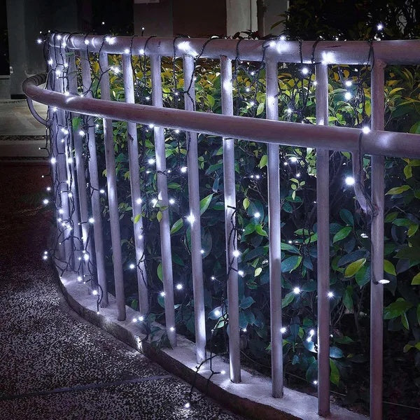 12m LED solar fairy string lights in cool white glowing on a metal railing in front of green garden bushes.