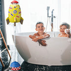 Yellow rocket sprinkler toy with space stickers, hovering by water pressure above kids in a white bathtub.