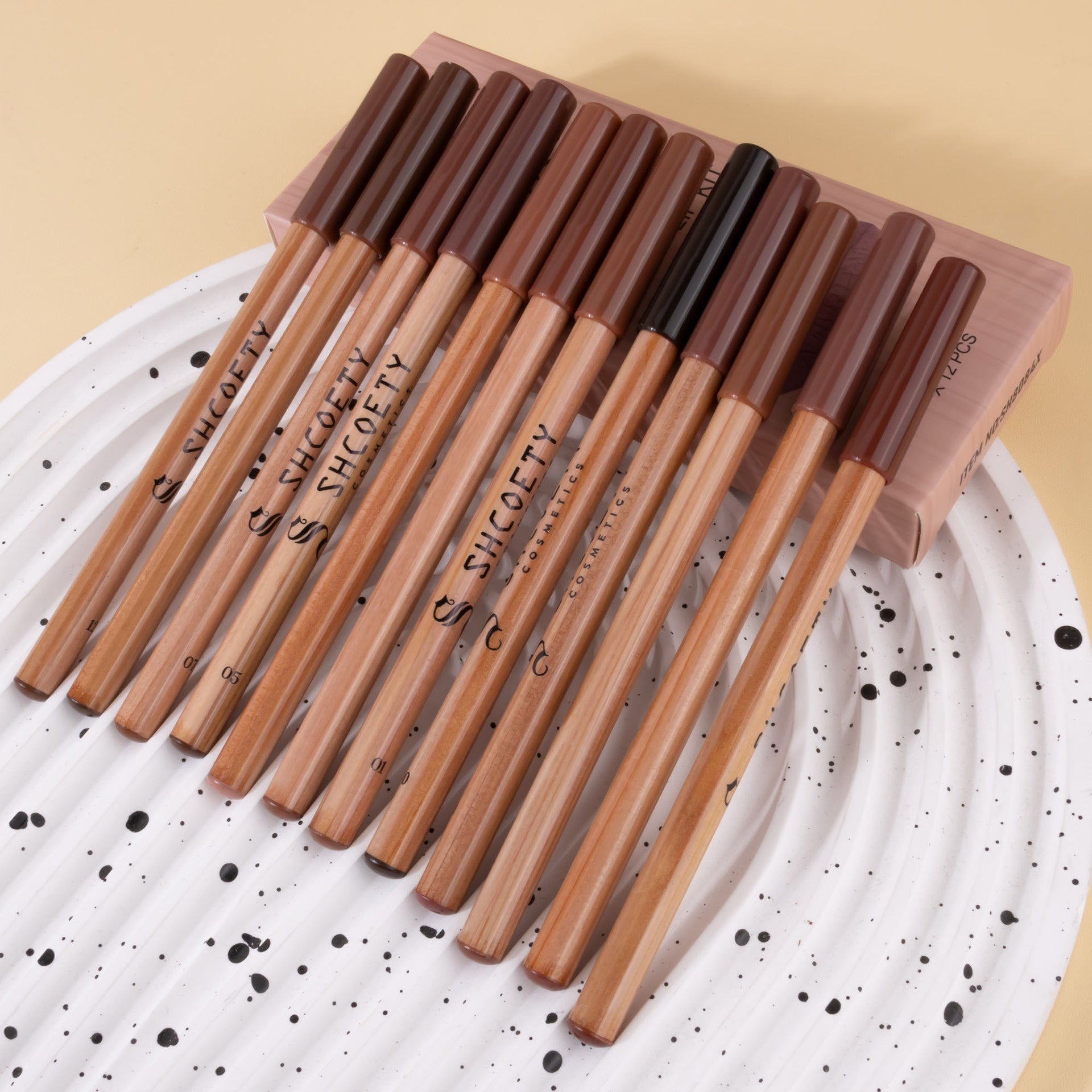 Twelve wooden-cased Shcoety matte lip liners in neutral shades, arranged on a white speckled plate in their box.