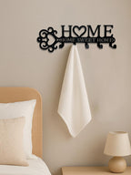 Black metal wall hook set featuring a "HOME" cutout with a heart and towel, mounted on a neutral bedroom wall.