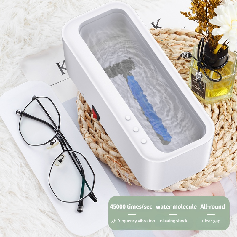 White ultrasonic cleaner with a blue-handled razor submerged in water, next to black-framed glasses on a woven mat.