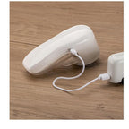 White electric lint remover with gold accent, connected by a USB cable to a power source on a wooden table.