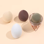 Four multi-colored makeup sponges, three loose and one held in a rose gold wire stand on a beige background.