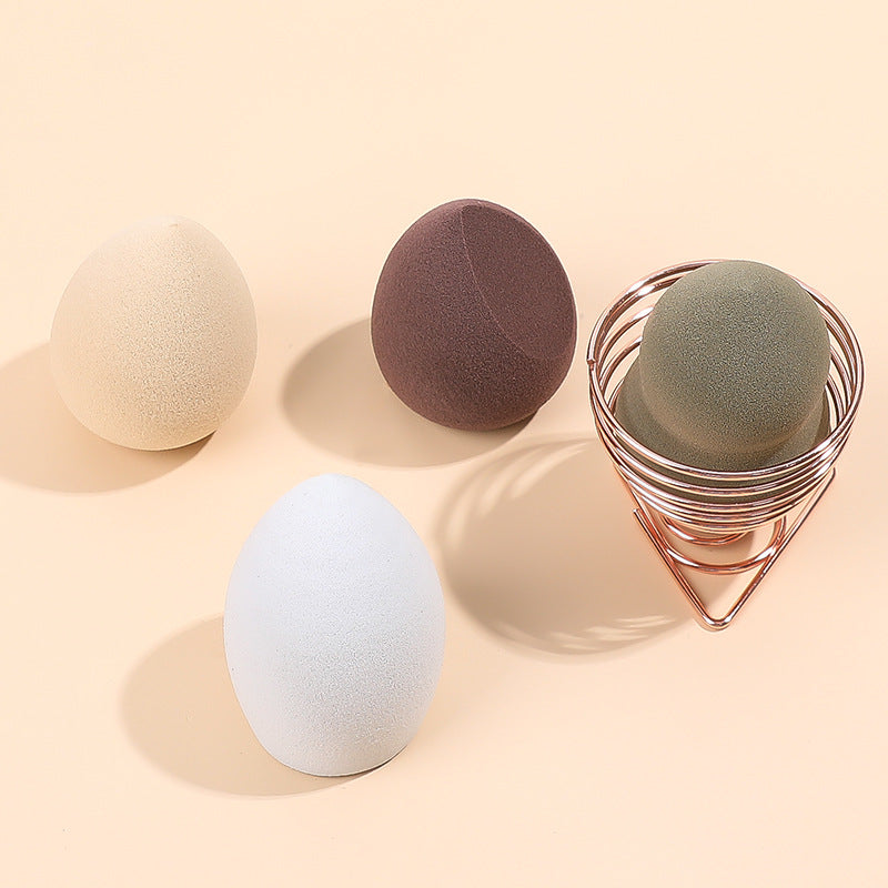 Four multi-colored makeup sponges, three loose and one held in a rose gold wire stand on a beige background.