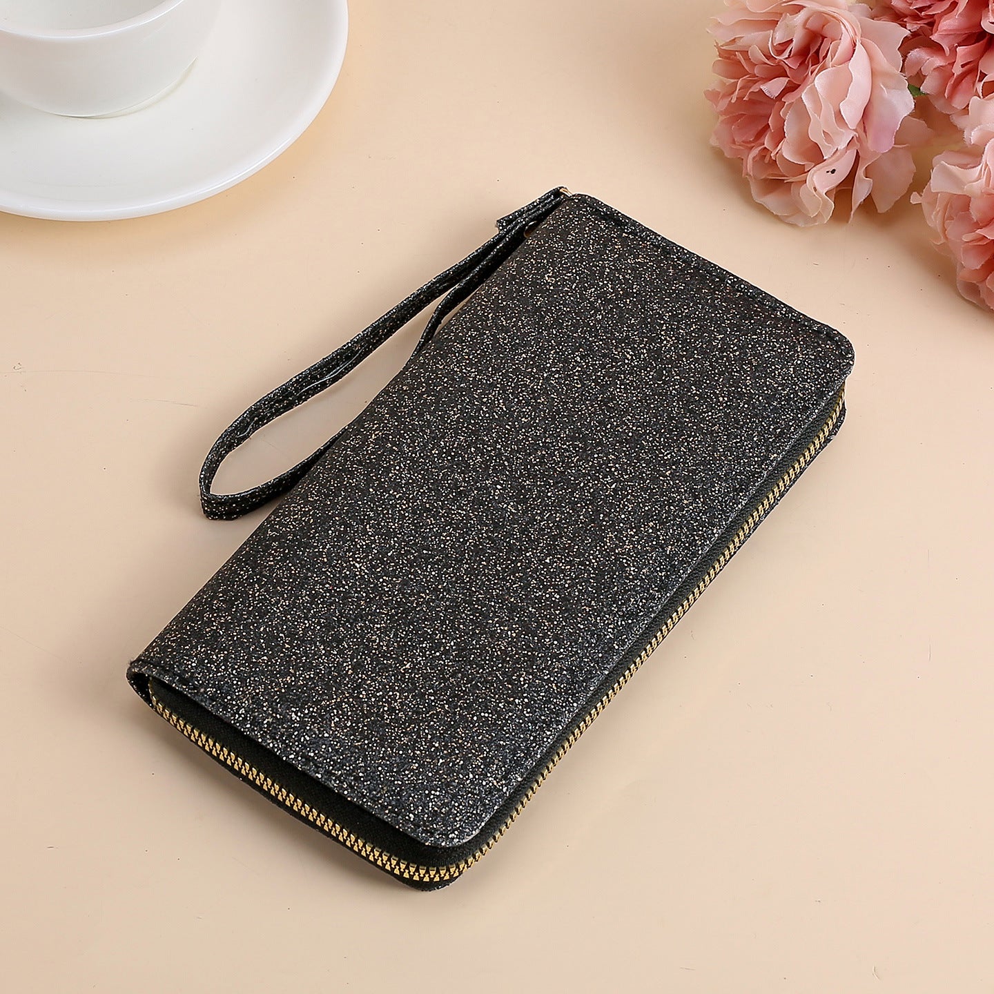 Black glittery rectangular clutch with a wrist strap and gold zipper, set on a table with a teacup and pink flowers.