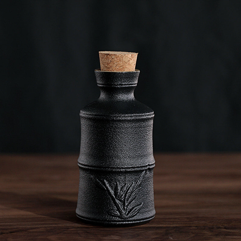Black, textured ceramic wine bottle with bamboo relief, topped with a wooden cork, on a wooden surface.
