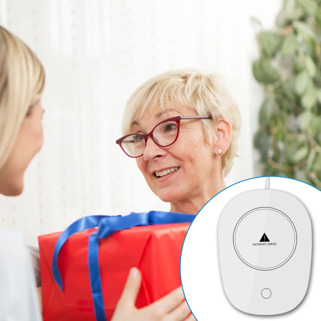 A white electric mug warmer with a "caution-hot surface" icon, shown alongside a person giving a gift.