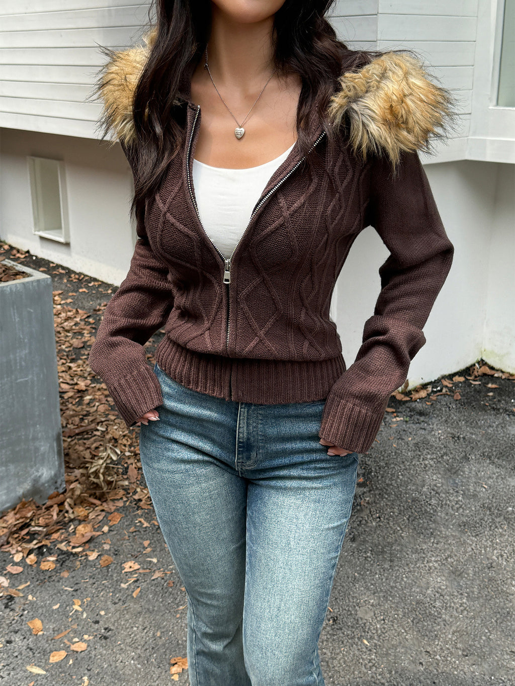 A brown, cable-knit, zip-up sweater coat featuring a faux fur-trimmed hood, worn over a white top and blue jeans.