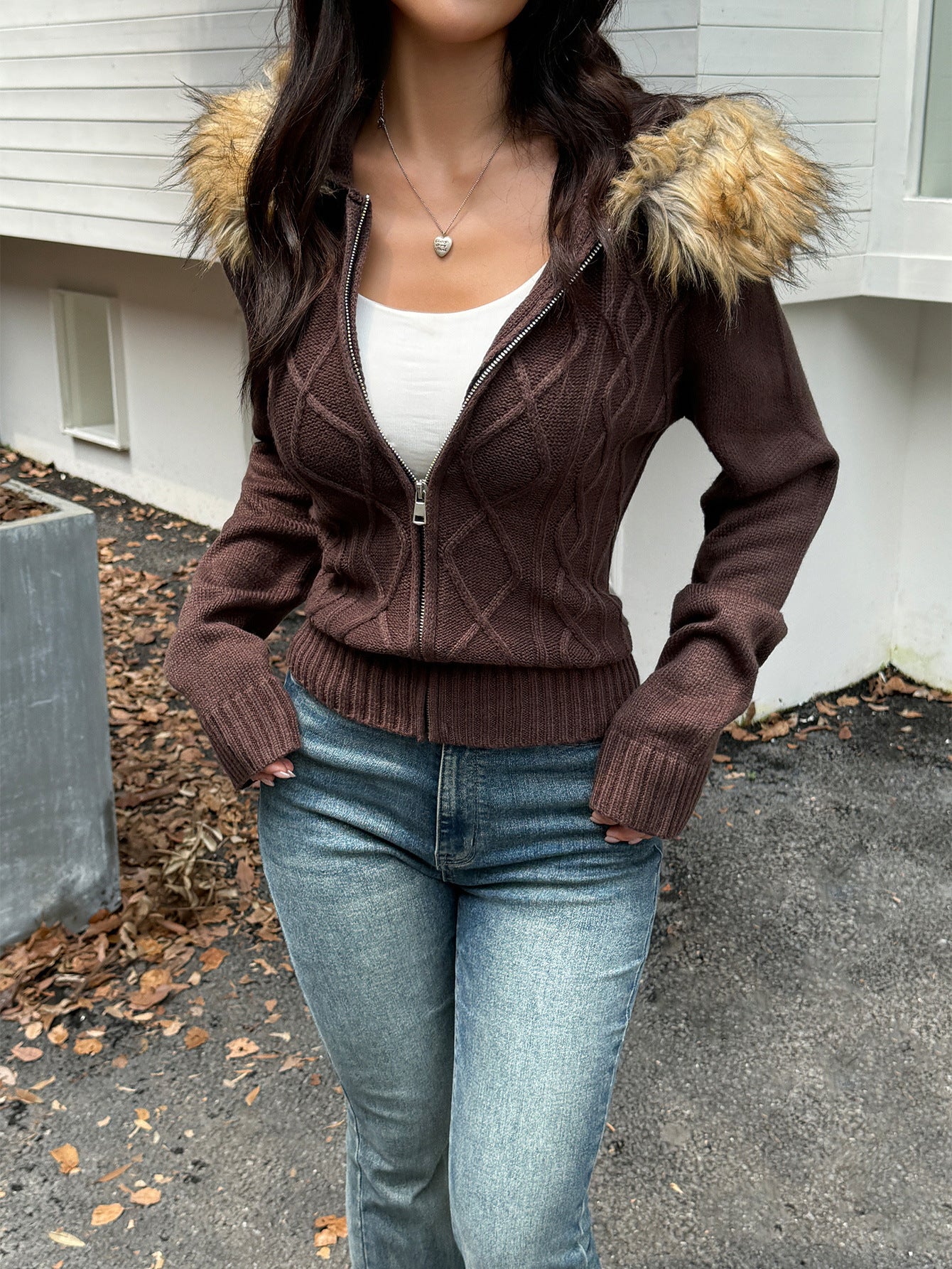 A brown, cable-knit, zip-up sweater coat featuring a faux fur-trimmed hood, worn over a white top and blue jeans.