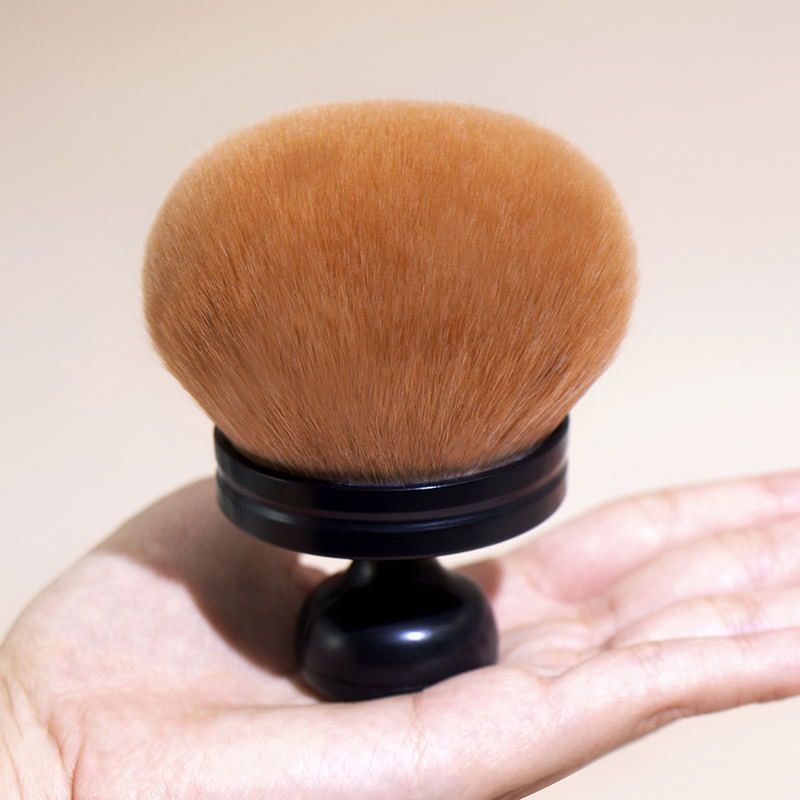 Handheld oversized makeup brush with a rounded tan bristle head and a sleek black handle, held in a palm.