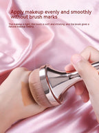Gold makeup brush with soft, dense bristles applying foundation to a hand against a pink satin background.