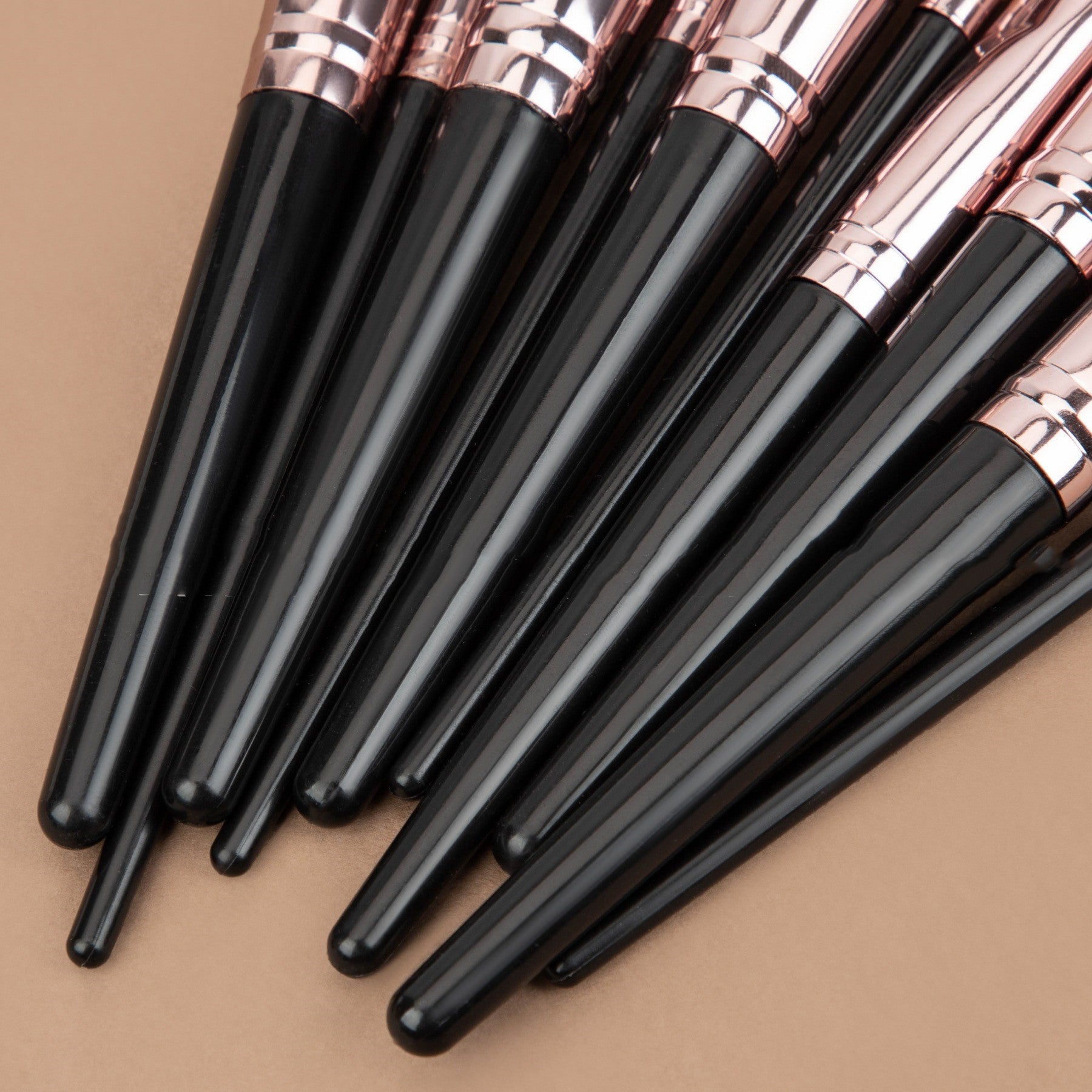 Set of makeup brushes with glossy black handles and metallic rose-gold ferrules on a tan background.