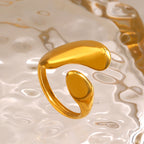 A minimalist gold-tone stainless steel open ring with fluid, rounded sculptural shapes on a rippled glass surface.