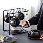 Matte black ceramic coffee cup set with gold trim on a sleek metal rack with matching saucers and spoons.