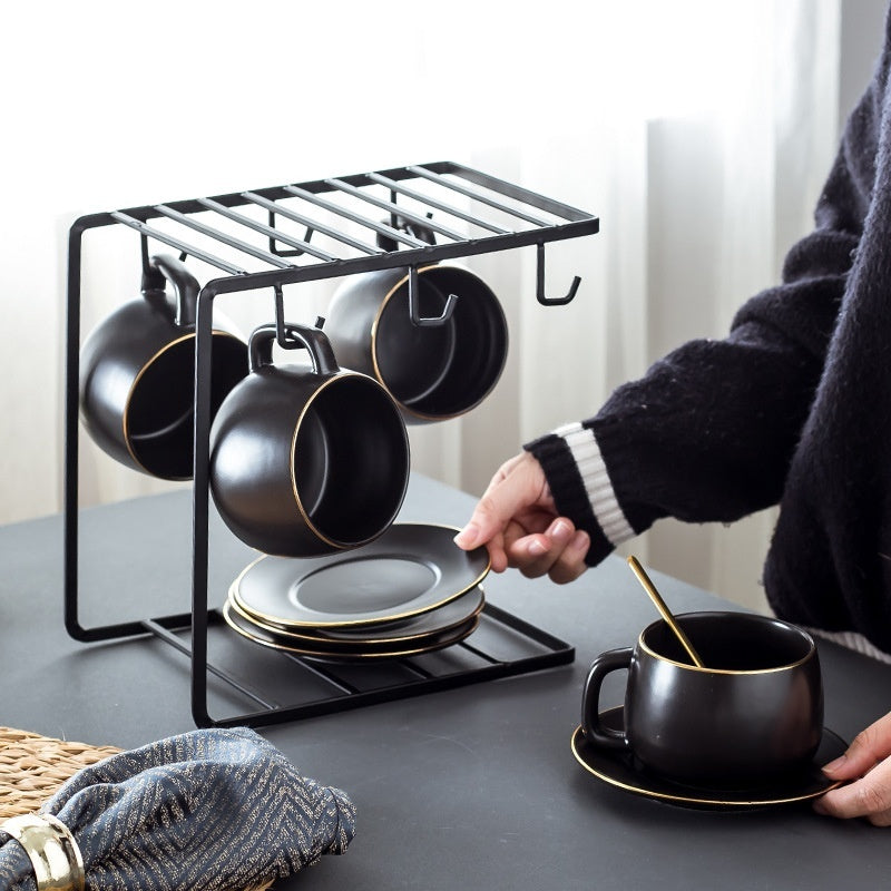 Matte black ceramic coffee cup set with gold trim on a sleek metal rack with matching saucers and spoons.