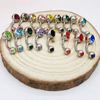 Assorted titanium steel belly button rings with colorful faceted crystal gems, arranged on a rustic wooden slice.