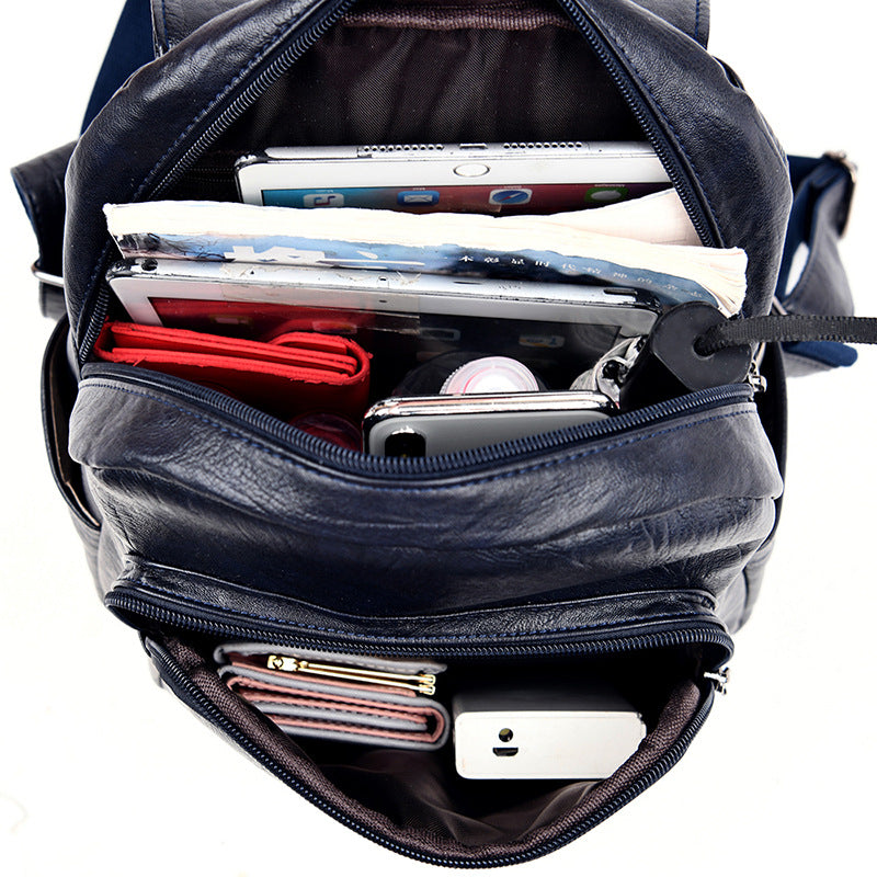 Open navy blue leather backpack filled with a tablet, book, wallet, and accessories, showing multiple zippered pockets.